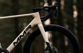 Primer plano de la mitad delantera de una bicicleta de carretera MMR Aelion SL blanca con frenos de disco y ruedas DT Swiss, aparcada en un sendero en un entorno forestal: una auténtica experiencia bici total.