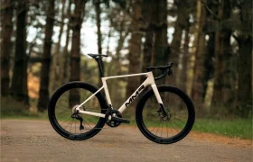 Una bicicleta de carretera MMR Aelion blanca con ruedas negras y manillar caído está aparcada en un camino pavimentado en una zona boscosa, encarnando el espíritu de bici total.
