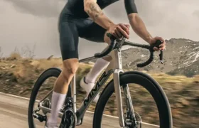 Un ciclista con ropa oscura y casco monta en una bicicleta de carretera MMR Aelion en un sendero al aire libre con montañas y cielos nublados de fondo.