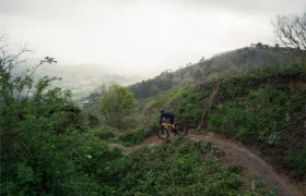 Una persona monta en bicicleta por un sinuoso sendero de tierra a través de un terreno verde y montañoso en un día nublado, mostrando una evolución total en su recorrido ciclista.