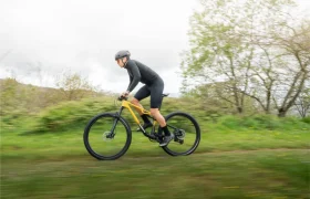 Una persona con casco y equipo de ciclismo monta una bicicleta de montaña amarilla MMR Kenta en un sendero cubierto de hierba, mostrando la evolución total, con árboles y un cielo nublado en el fondo.