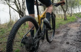 Un ciclista vestido de negro conduce una bicicleta de montaña amarilla MMR Kenta por un sendero fangoso y boscoso en un día nublado, mostrando una evolución total.
