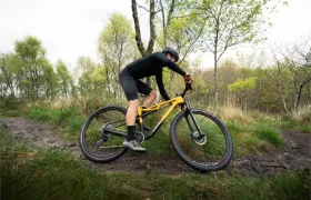 Vestido con equipo de ciclismo negro, un ciclista muestra su evolución total mientras aborda un sendero cuesta arriba fangoso en una MMR Kenta amarilla, rodeado de árboles y césped.