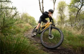 Una persona que usa casco conduce una bicicleta de montaña amarilla MMR Kenta en un sendero de tierra, mostrando la evolución total a través de un área boscosa y cubierta de hierba.