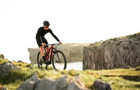 Una persona con equipo de ciclismo y casco conduce una bicicleta de montaña MMR Kenta, mostrando la evolución total en un terreno cubierto de hierba y rocoso cerca de un acantilado con agua y más acantilados detrás.