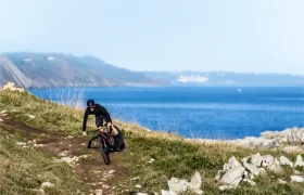 Un ciclista vestido de negro recorre una bicicleta de montaña MMR Kenta por un sendero costero rocoso, capturando la esencia de evolución total con el mar y los acantilados distantes visibles en el fondo.