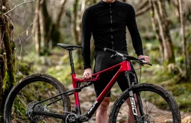 Un ciclista con equipo y casco negros se encuentra al aire libre en un sendero rocoso, sosteniendo una bicicleta de montaña MMR Kenta roja y negra, lista para la evolución total en el área boscosa.