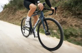 Un ciclista vestido de negro conduce una bicicleta de ruta azul MMR Simun por un camino pavimentado, rodeado de tierra y vegetación.