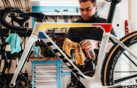 En su taller, un hombre utiliza el spray limpiador Motul Bike Care y una toalla para limpiar el cuadro de una bicicleta de carretera, rodeado de herramientas y equipamiento Bicimax.