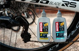Dos botellas de lubricante de cadena Motul Bike Care y Bicimax están colocadas en posición vertical en el suelo junto a la rueda trasera y el descarrilador de una bicicleta.