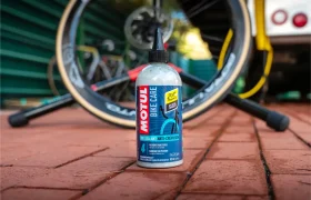 Una botella de sellador para bicicletas Motul, de la gama Motul Bike Care, reposa sobre una superficie de ladrillo, con una rueda de bicicleta y otra bicicleta Bicimax difuminadas en el fondo.