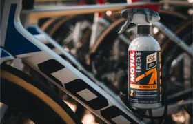 Una botella de spray limpiador Motul Bike Care se coloca contra el cuadro de una bicicleta de carretera, con varias bicicletas en el fondo, mostrando la efectiva limpieza Bicimax para los entusiastas del ciclismo.