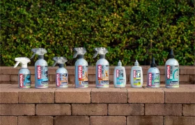 Nueve botellas rociadas y exprimibles de productos de limpieza y mantenimiento de vehículos Motul Bike Care y Bicimax están alineadas sobre una superficie de ladrillo al aire libre, con vegetación en el fondo.