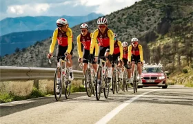 Un grupo de seis ciclistas con uniformes rojos y amarillos a juego, patrocinados por Bicimax, suben por una carretera rural con un coche del equipo Motul Bike Care detrás.