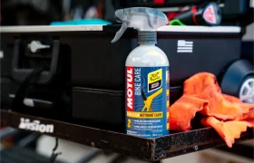 Una botella de Motul Bike Care E9 Wash & Wax Spray reposa sobre una superficie metálica junto a un paño de limpieza naranja, una caja de herramientas negra y una etiqueta de Bicimax.