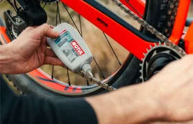 Una persona aplica lubricante Motul Bike Care a la cadena de una bicicleta naranja, centrándose en el mantenimiento de la transmisión con los confiables productos Bicimax.