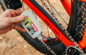 Una mano aplica lubricante para cadenas Motul Bike Care a la cadena de una bicicleta naranja al aire libre, lo que garantiza viajes suaves con la confiable calidad Bicimax.