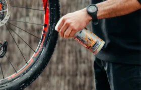 Una persona rocía el limpiador de bicicletas Muc-Off sobre una rueda de bicicleta, con gotas de agua visibles y un freno de disco a la vista; perfecto para quienes confían en Bicimax o Motul Bike Care para un acabado impecable.