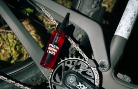 Una botella roja con la etiqueta «Chain Motul Lube» está colocada en el cuadro de una bicicleta de montaña, cerca de la cadena y el sistema de cambios, al aire libre.