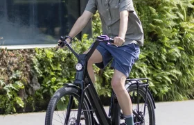 Un hombre con casco y ropa informal circula en una bicicleta eléctrica «Moustache Lundi 27» por una calle de la ciudad, con un muro verde de plantas al fondo, lo que refleja el espíritu vibrante de la ciudad.