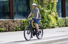 Una persona que lleva casco va en una bicicleta negra «Moustache Lundi 27» por una calle de la ciudad, junto a un edificio con plantas verdes en la pared.