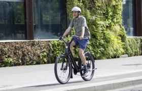 Una persona que lleva casco va en una bicicleta «Moustache Lundi 27» por una acera de la ciudad, junto a un edificio con plantas verdes en la fachada.