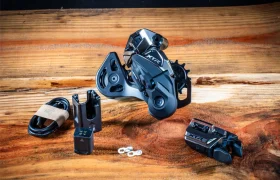 Cambio trasero electrónico Shimano XTR Di2, palanca de cambios, cable de carga y pequeñas piezas de ferretería de los kits de actualización de Shimano dispuestos sobre una superficie de madera.