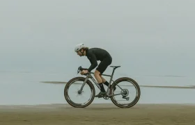 Una persona que viste un casco y ropa de ciclismo MMR Simun conduce una bicicleta de carretera por un paisaje llano y arenoso cerca del agua bajo un cielo nublado.