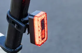 Un piloto trasero rojo Sigma Nyte 70 se sujeta a la tija del sillín de una bicicleta con una correa negra, sobre un fondo gris liso.