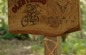 Cartel de madera situado en la ruta de BTT de Olentzero en el que aparecen un hombre en bicicleta, el símbolo de un ciervo y el texto «Trail Born» junto al logotipo de Shimano.