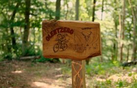 Señal de madera para la red de rutas de BTT de Basaburua con una ilustración de un ciclista de montaña, el texto «Olentzero» y un logotipo con cuernos de ciervo.