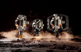 Tres estilos diferentes de pedales de bicicleta, incluidos los pedales para Gravel del ecosistema de pedales LOOK, se exponen en posición vertical sobre una superficie texturada y polvorienta, con una iluminación espectacular y un fondo oscuro.