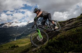Un ciclista de montaña, equipado con protecciones, desciende por un sendero rocoso, confiando en los neumáticos Pirelli Scorpion MTB para obtener un agarre y un control óptimos, con las montañas y las nubes como telón de fondo.