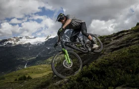 Un ciclista de montaña desciende por un sendero rocoso, equipado con neumáticos Pirelli Scorpion para MTB, rodeado de verdes colinas y lejanas montañas nevadas bajo un cielo nublado.