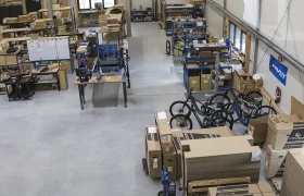 Vista aérea de la inauguración de un taller de montaje de bicicletas: diversos puestos de trabajo, piezas en cajas y bicicletas parcialmente montadas repartidas por el suelo de hormigón en previsión de convertirse en el nuevo pivote de operaciones de su sede central europea.