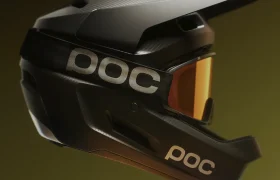 Un casco integral POC negro con visera y gafas de color naranja, perfecto para descensos, sobre un fondo amarillo oscuro con degradado.