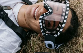 Una persona con camisa blanca y gafas de esquí Ora yace sonriente sobre hierba seca, con un casco negro Coron Air rotulado "POC" cerca.