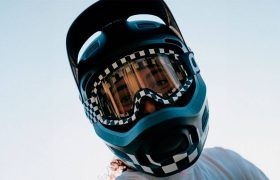 Persona vestida con un casco Coron Air negro con detalles a cuadros y gafas transparentes Ora, mirando hacia la cámara sobre un fondo de cielo despejado, que recuerda el atrevido estilo de motocross de Robin Goomes.