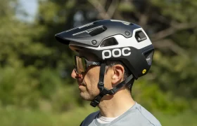 Un hombre que lleva un casco de ciclista POC Cularis Pure blanco y negro, con visera y gafas de sol, está de pie al aire libre, preparado para protegerse del sol, con un fondo verde difuminado.
