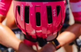 Un ciclista con casco y maillot rosa se inclina hacia delante sobre el manillar, con las manos entrelazadas en posición de carrera, manteniéndose fresco con un casco ventilado durante el calor extremo.