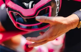 Una persona con ropa de ciclismo sostiene un casco ventilado rosa y negro sobre una bicicleta, con fondo borroso.