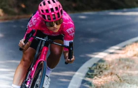 Un ciclista con una equipación rosa y casco ventilado monta en una bicicleta de carretera rosa, inclinándose en una curva de una carretera asfaltada rodeada de árboles y hierba seca.