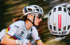 Un ciclista que lleva casco y gafas de sol pedalea a toda velocidad; a la derecha se ve un primer plano del casco blanco con pegatinas y texto.