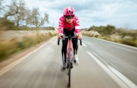 Un ciclista que viste un casco POC Procen Air rosa y una camiseta a juego monta en bicicleta con un efecto de movimiento borroso contra un cielo nublado, encarnando el espíritu de EF Pro Cycling.