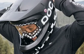 Una persona con camiseta negra de manga larga se ajusta un casco integral negro Coron Air POC con gafas Ora, al aire libre cerca de una zona boscosa.
