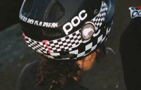 Una persona lleva un casco negro de ciclista con un estampado de damero, el logotipo "POC" y el texto "EVERY DAY IS A DREAM" en la parte trasera, similar al icónico estilo de Robin Goomes y su casco favorito Coron Air.