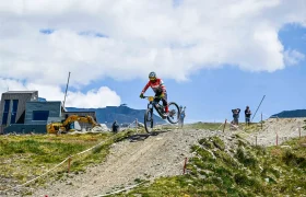 Un ciclista de montaña, con todo el equipo puesto, salta por encima de una rampa de tierra en un sendero al aire libre, mientras los espectadores observan y se ve un vehículo de obra al fondo.