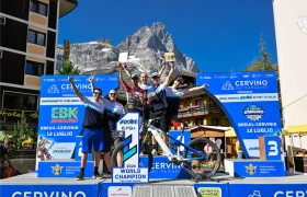 Cinco ciclistas posan en un podio con trofeos y medallas en la mano, con una montaña y pancartas del evento al fondo, en un campeonato de ciclismo.