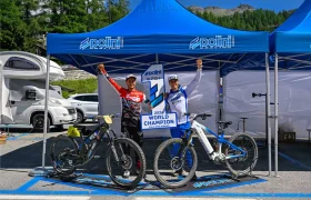 Dos ciclistas con equipación de competición están de pie junto a sus bicicletas bajo una carpa azul, sosteniendo un cartel que dice «Campeón del Mundo 2024» y levantando los brazos en señal de celebración.