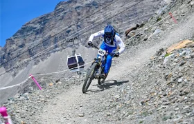 Un ciclista de montaña, vestido con ropa azul y blanca, desciende por un sendero rocoso; al fondo se ve una placa con el número 401 y un teleférico.